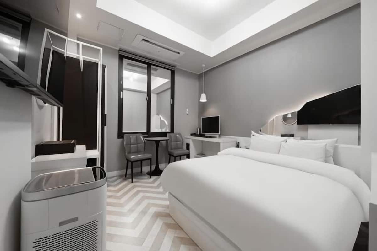 A modern hotel room with a large bed, a desk, a chair, a TV, and a trash bin.