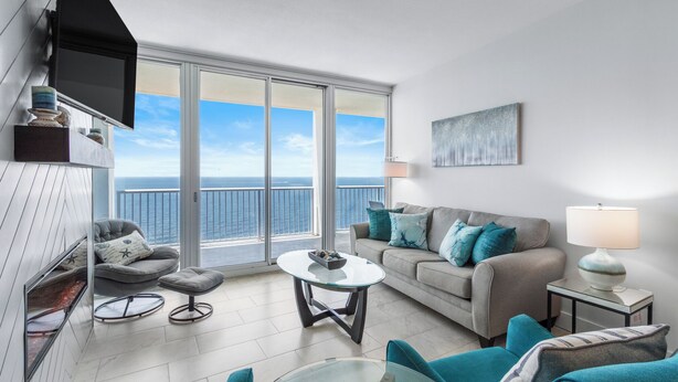 Ein modernes Wohnzimmer mit Balkon und Meerblick, ein graues Sofa mit türkisfarbenen Kissen, ein kleiner runder Tisch und ein an der Wand befestigter Fernseher.
