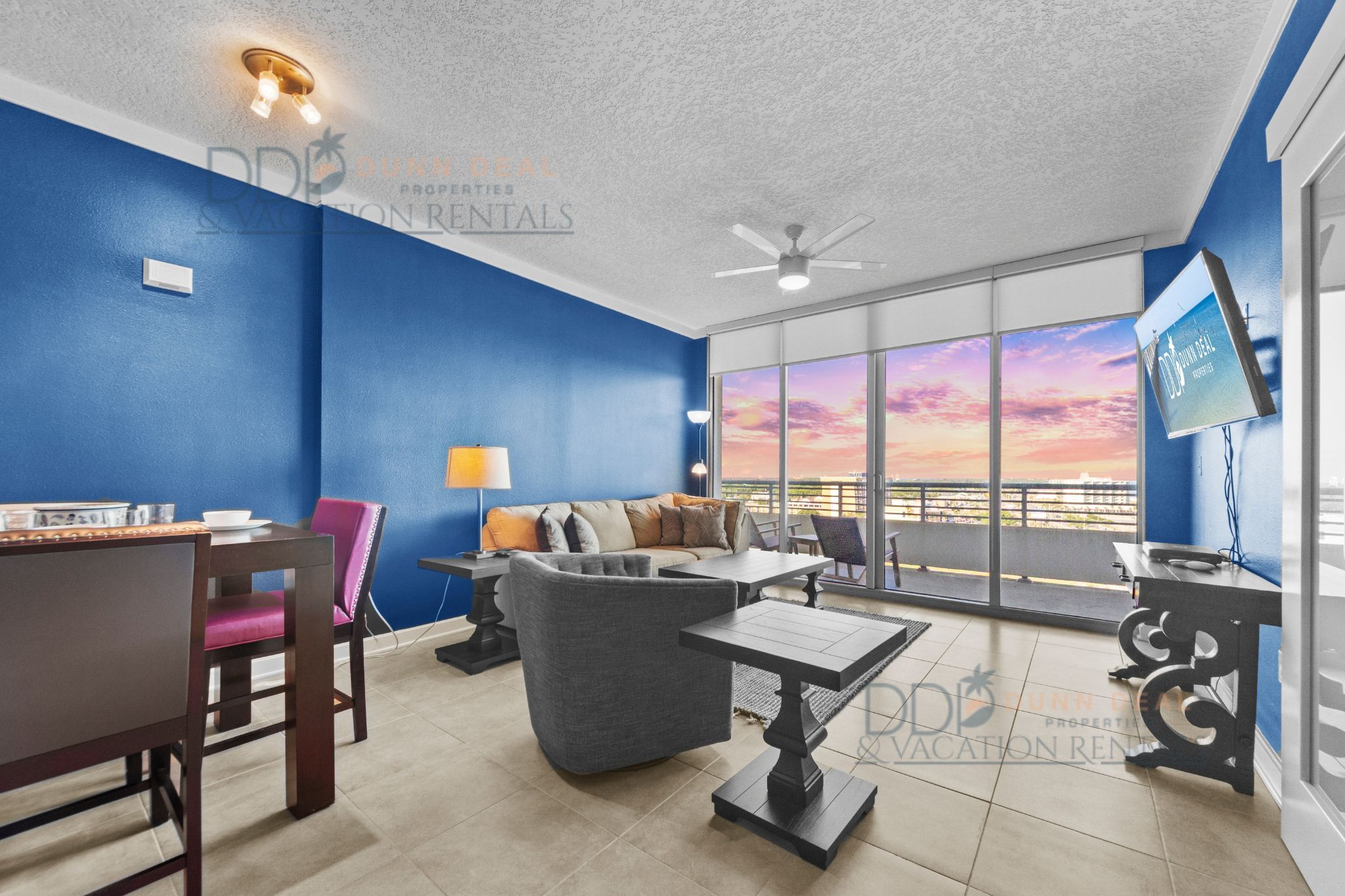 Un salon moderne avec un mur d’accent bleu, un canapé, une table à manger avec des chaises, un téléviseur et un balcon offrant une vue sur le coucher de soleil.