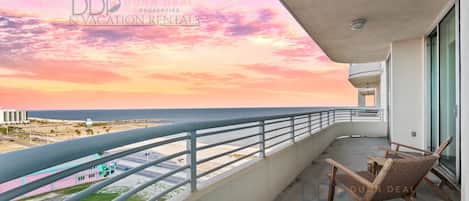 A balcony with a view of the ocean and beach, wooden chairs, and a railing.