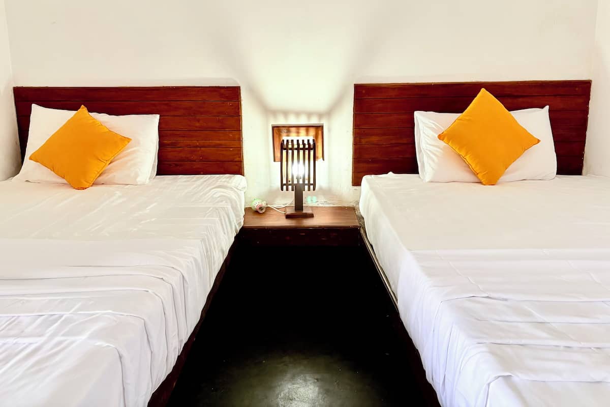 A hotel room with two single beds, each with white bedding and a wooden headboard. There is a nightstand with a lamp between the beds.