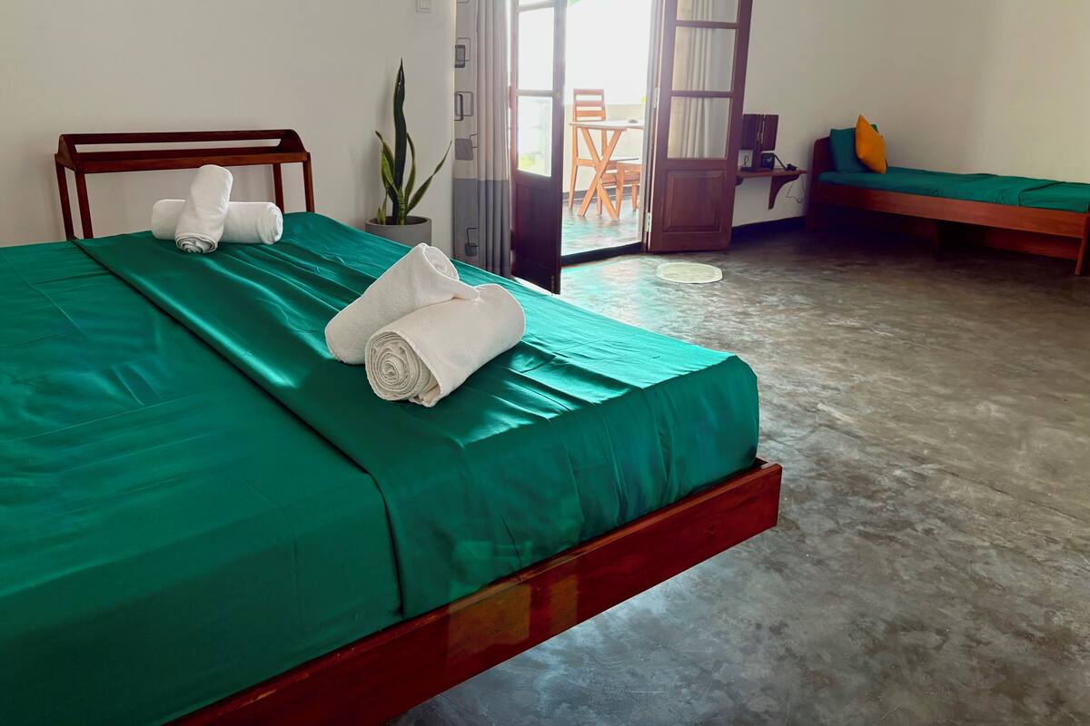 A neatly made bed with green sheets and two rolled towels, a wooden headboard, a potted plant, and a view of the outside through an open door.