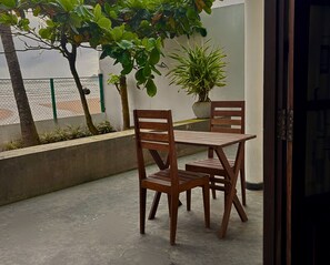A wooden table and chairs on a balcony with a concrete floor, surrounded by greenery and a view of the sea.