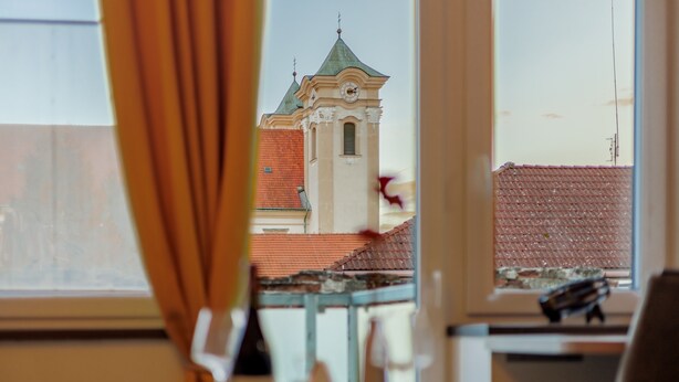 Vista a través de una ventana con cortina, un edificio con una torre de reloj y un tejado rojo.