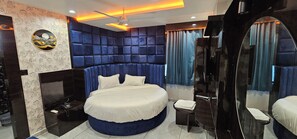 A modern bedroom with a large bed, a built-in headboard, a bedside table, a mirror, and a ceiling fan. - hotel shagun inn bopal (Ahmedabad)