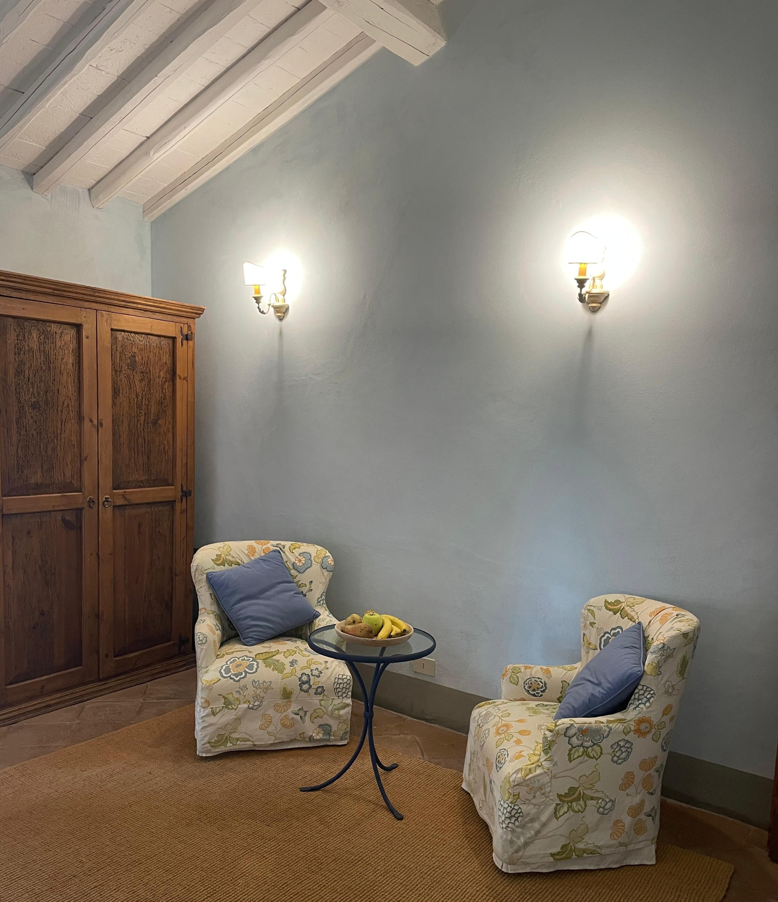 Une pièce comprenant deux fauteuils à motifs floraux, une petite table ronde avec des fruits et une armoire en bois.