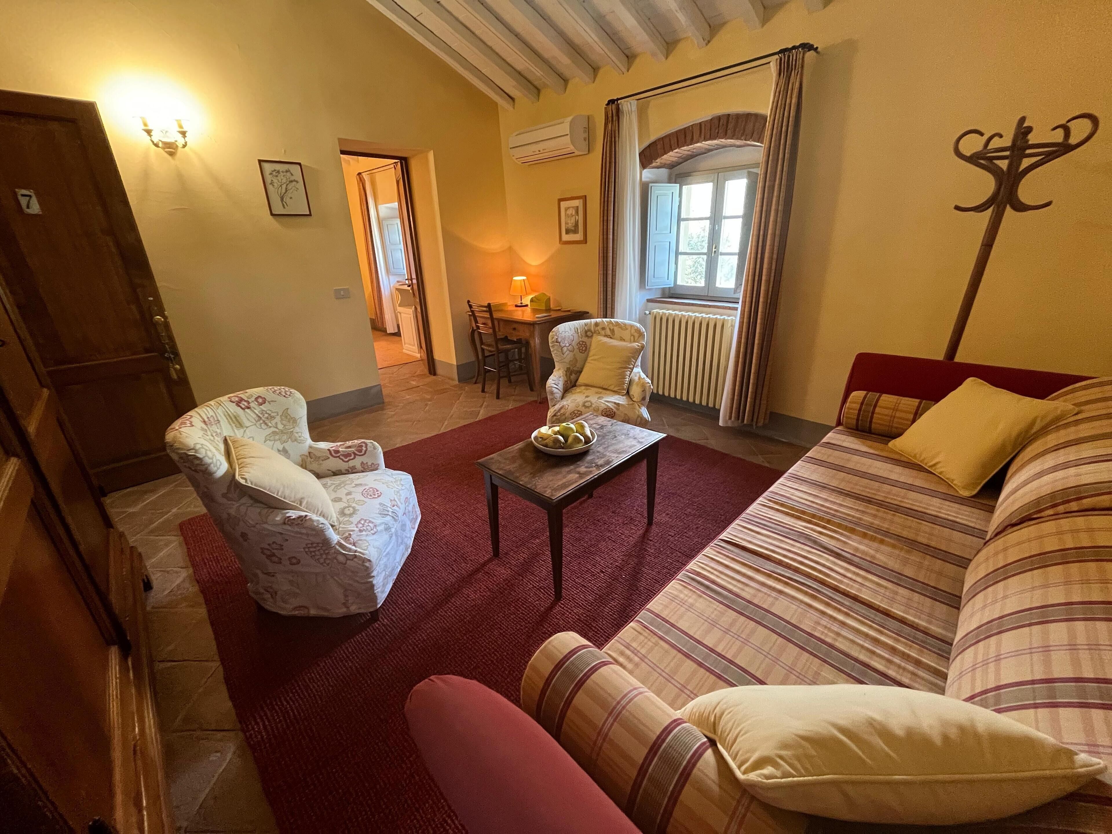A cozy living room with a sofa, armchair, and a small table with fruit. There is a window with curtains, a radiator, and a door leading to another room.