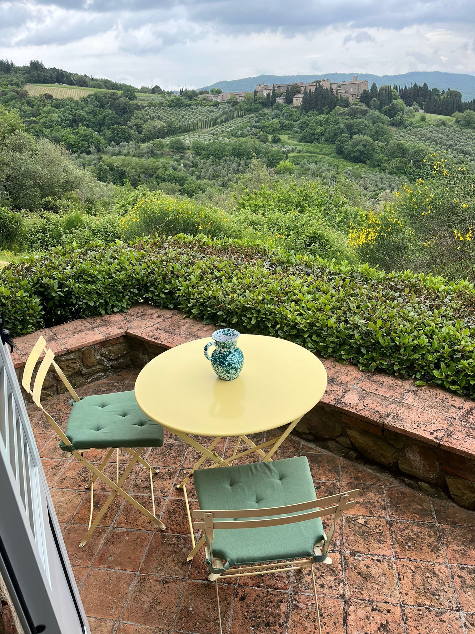 Une terrasse avec une table ronde jaune et deux chaises vertes à coussins, d’où l’on peut admirer un paysage luxuriant avec des collines et, au loin, un bâtiment historique.