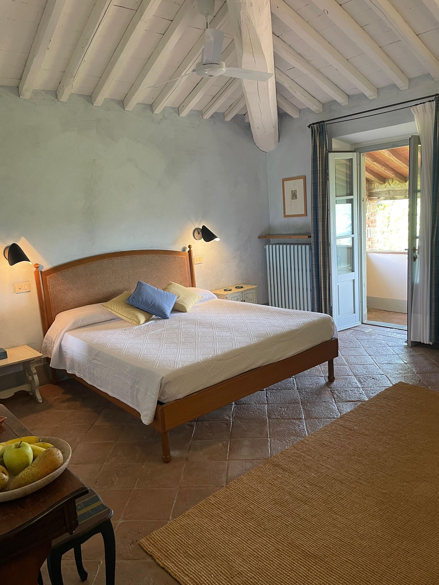 Un dormitorio con cama de madera, una mesita de noche, lámpara, radiador y vistas al exterior a través de un ventanal.