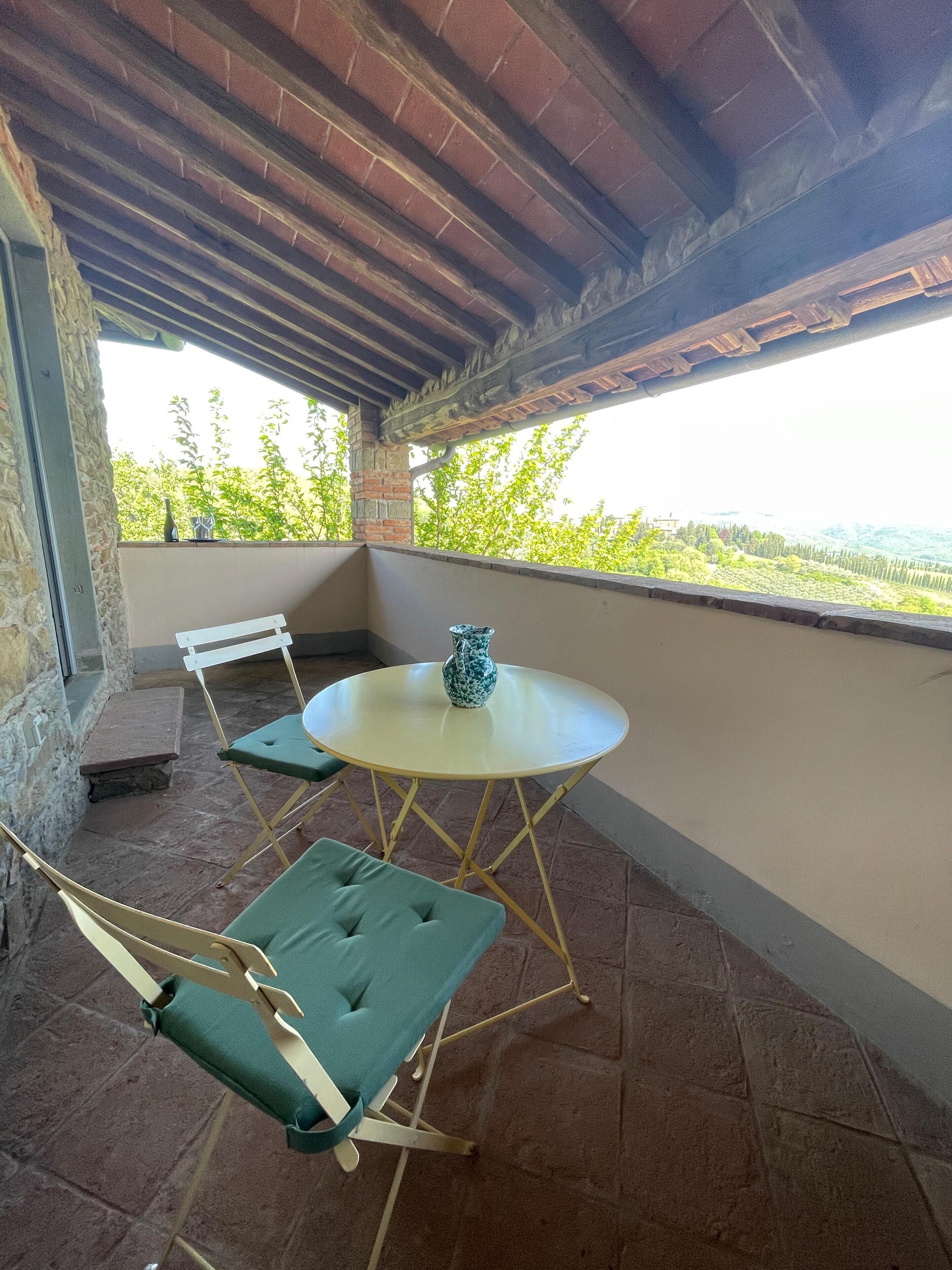 A covered patio with a round table and two chairs, a stone bench, and a view of greenery and hills.