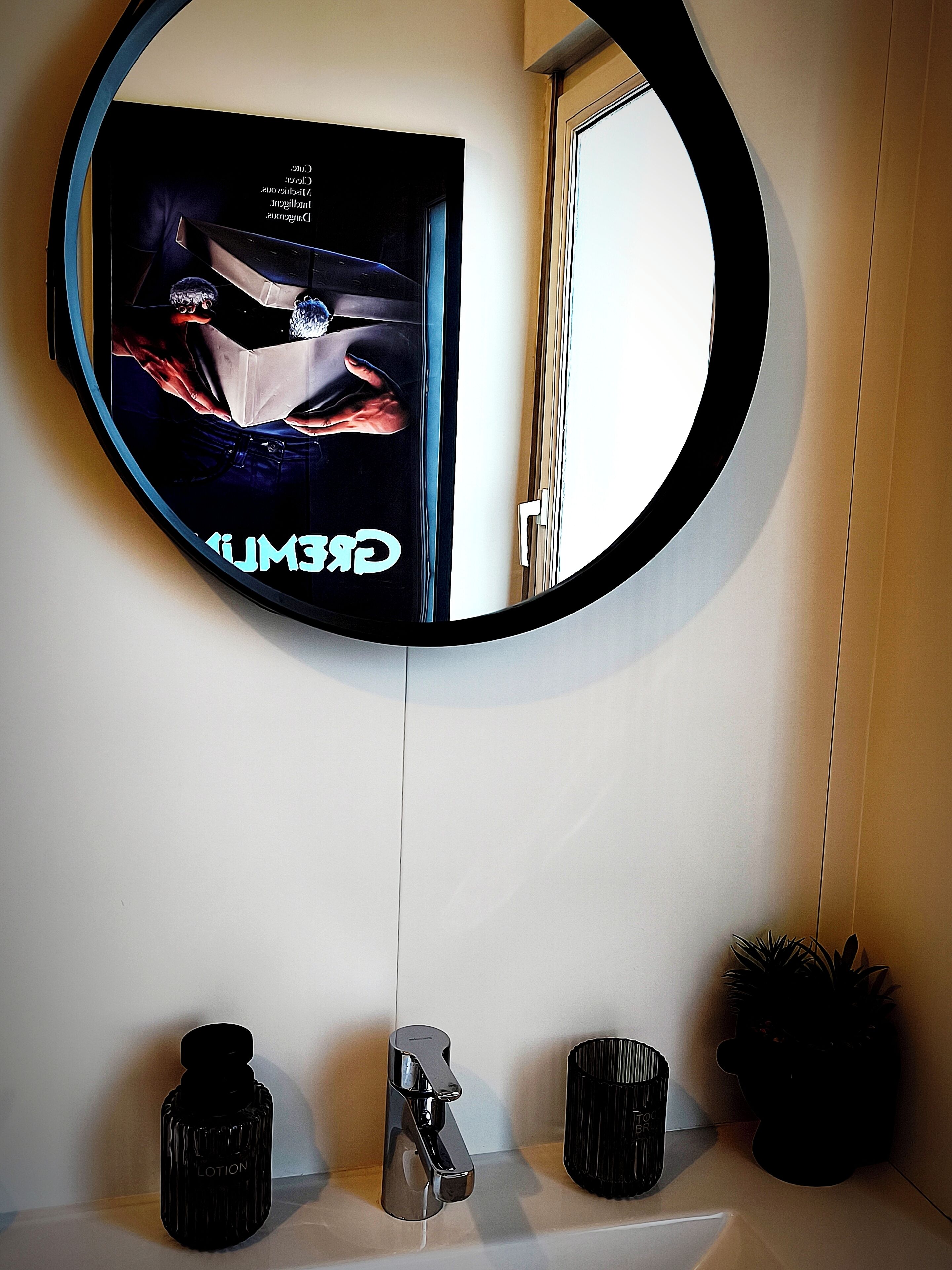 Een badkamer met een ronde spiegel, een wastafel, een kraan, een fles met de tekst 'LOTION' en een afvalbak.
