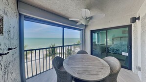 A balcony with a wooden table and wicker chairs, overlooking a beach. - Seaside Resort 507 | Fresh Renovation + Beachfront Living (Fort Myers Beach)