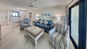 A modern living room with a sofa, armchairs, and a coffee table. There is a kitchen in the background with white cabinets and a window. - Seaside Resort 507 | Fresh Renovation + Beachfront Living (Fort Myers Beach)