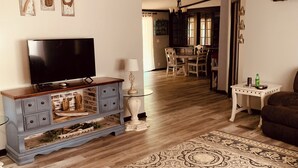 A living room with a television, a ceiling fan, a patterned rug, and a dining area in the background.