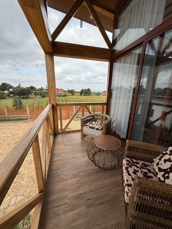 A wooden balcony with wicker furniture, a stone floor, and a view of a field and houses in the distance.