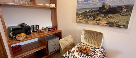A compact kitchen with wooden shelves, a microwave, and a small refrigerator. There is a wicker chair with a patterned cushion and a table with a decorative cloth. A landscape painting adorns the wall.