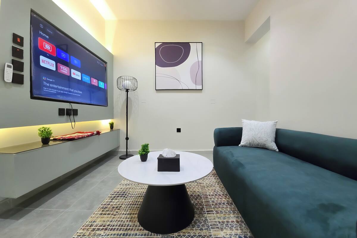 Ein modernes Wohnzimmer mit einem Wand-Fernseher, einem runden Couchtisch, einem grünen Sofa und einem gemusterten Teppich.