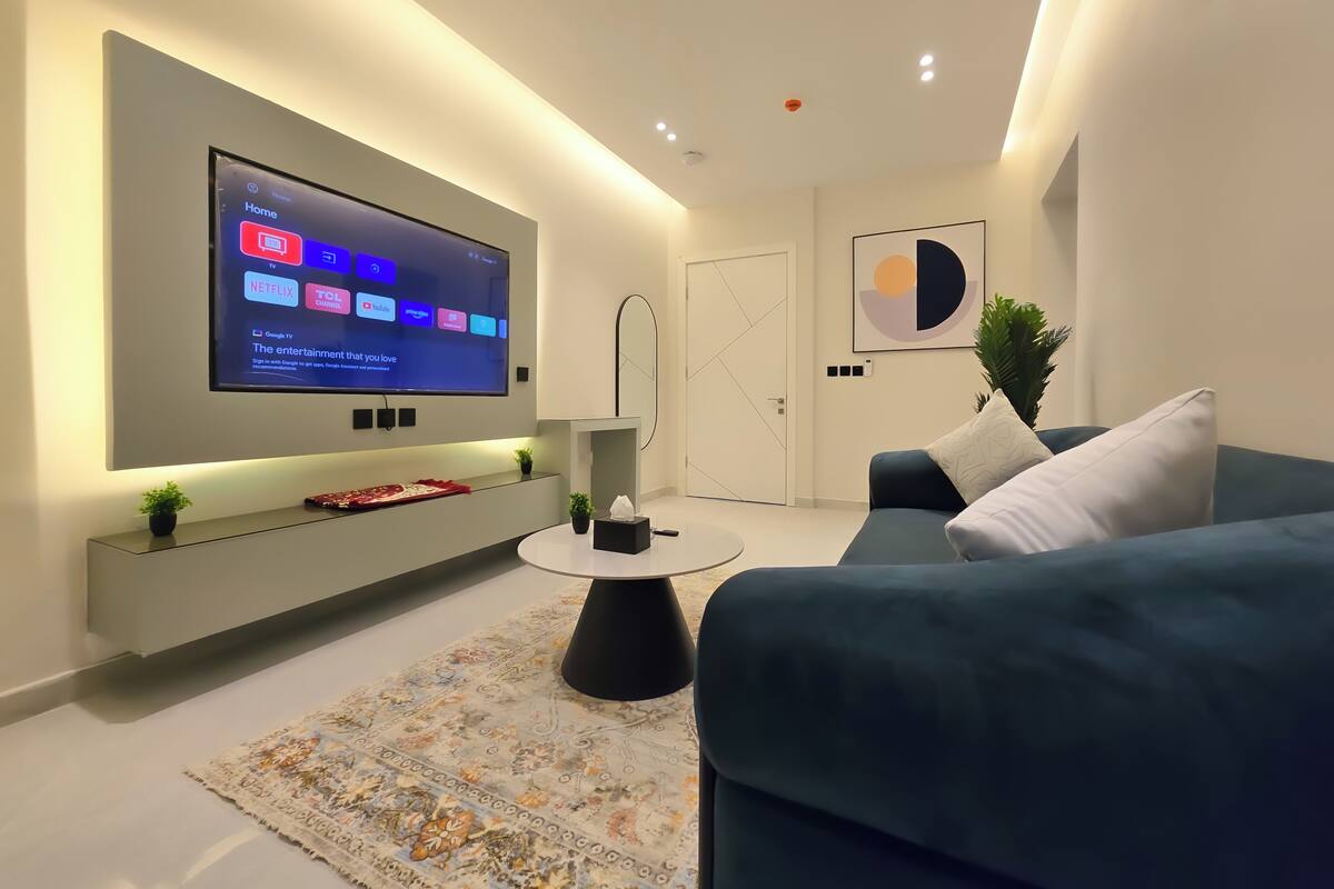 Ein modernes Wohnzimmer mit einem an der Wand befestigten Flachbildfernseher, einem blauen Sofa, einem kleinen Beistelltisch und einem gemusterten Teppich.