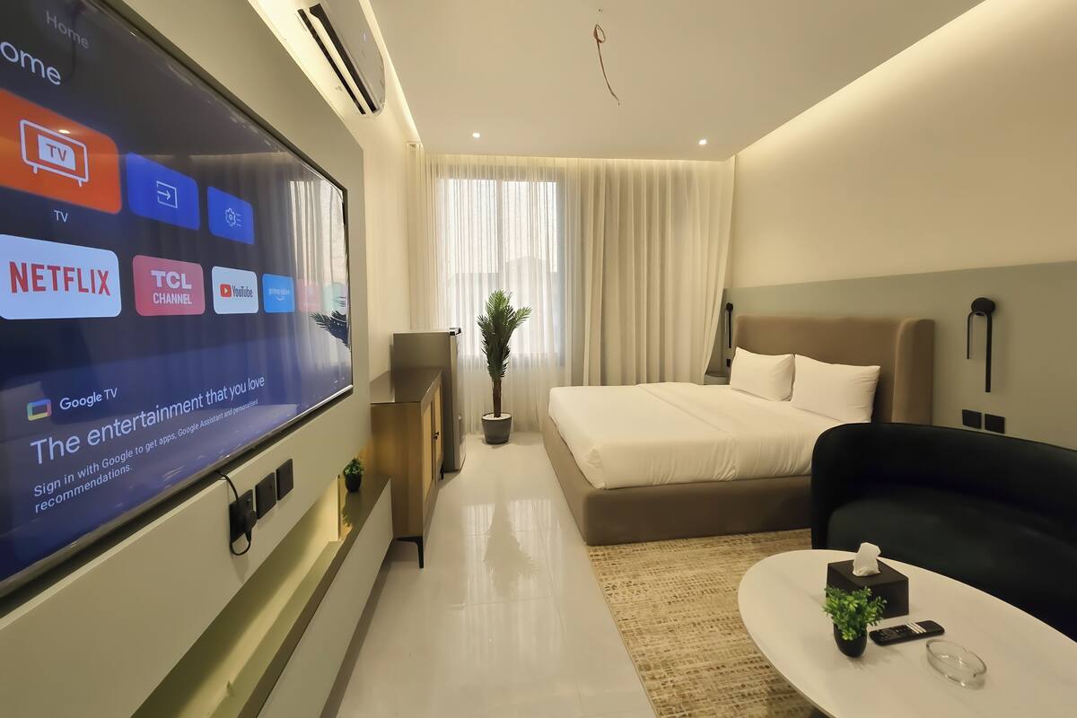 Ein modernes Hotelzimmer mit großem Fernseher, der Streaming-Dienste anzeigt, einem Bett, einer Couch, einem kleinen Tisch mit Pflanze und einem Nachttisch mit Fernbedienung.
