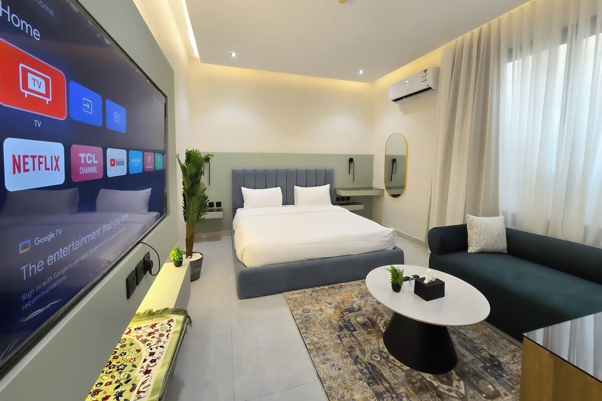 Ein modernes Hotelzimmer mit einem großen Fernseher, der verschiedene Streaming-Dienste anzeigt, einem ordentlich bezogenen Bett, einem Sofa, einem kleinen Couchtisch und einer Topfpflanze.