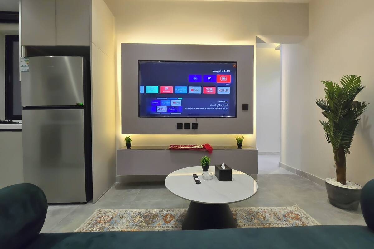 Eine moderne Küche mit integriertem Fernseher, der verschiedene Streaming-Dienste anzeigt, einem Kühlschrank und einem runden Esstisch mit einer kleinen Pflanze und Fernbedienungen.