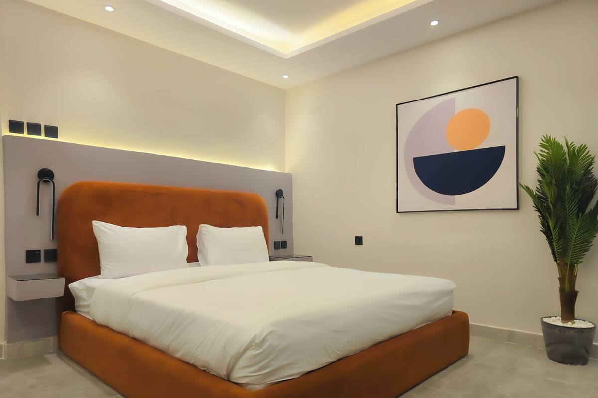 Ein modernes Schlafzimmer mit einem großen Bett, einem mit Beleuchtung versehenen Kopfteil, einem Nachttisch, einem an der Wand befestigten abstrakten Gemälde und einem Blumentopf.