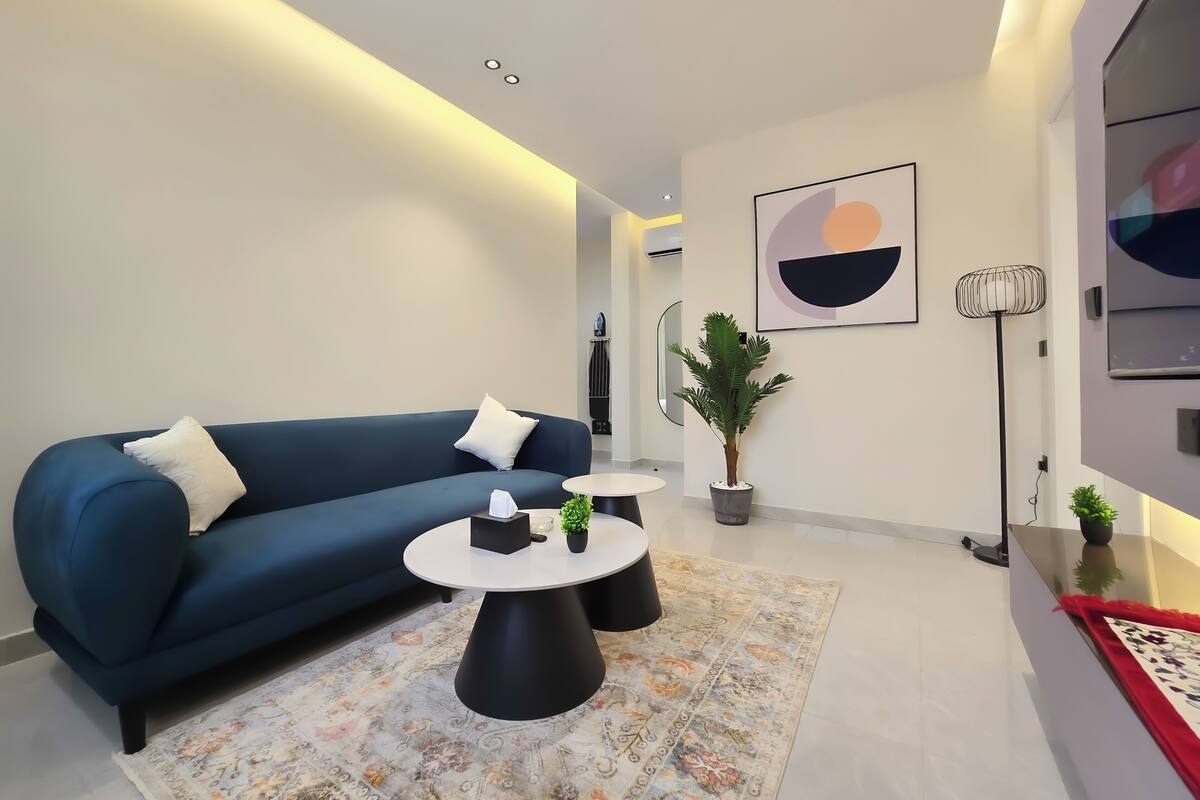 Ein modernes Wohnzimmer mit einem blauen Sofa, zwei schwarzen Couchtischen, einem gemusterten Teppich, einer Topfpflanze und einem Bild an der Wand.