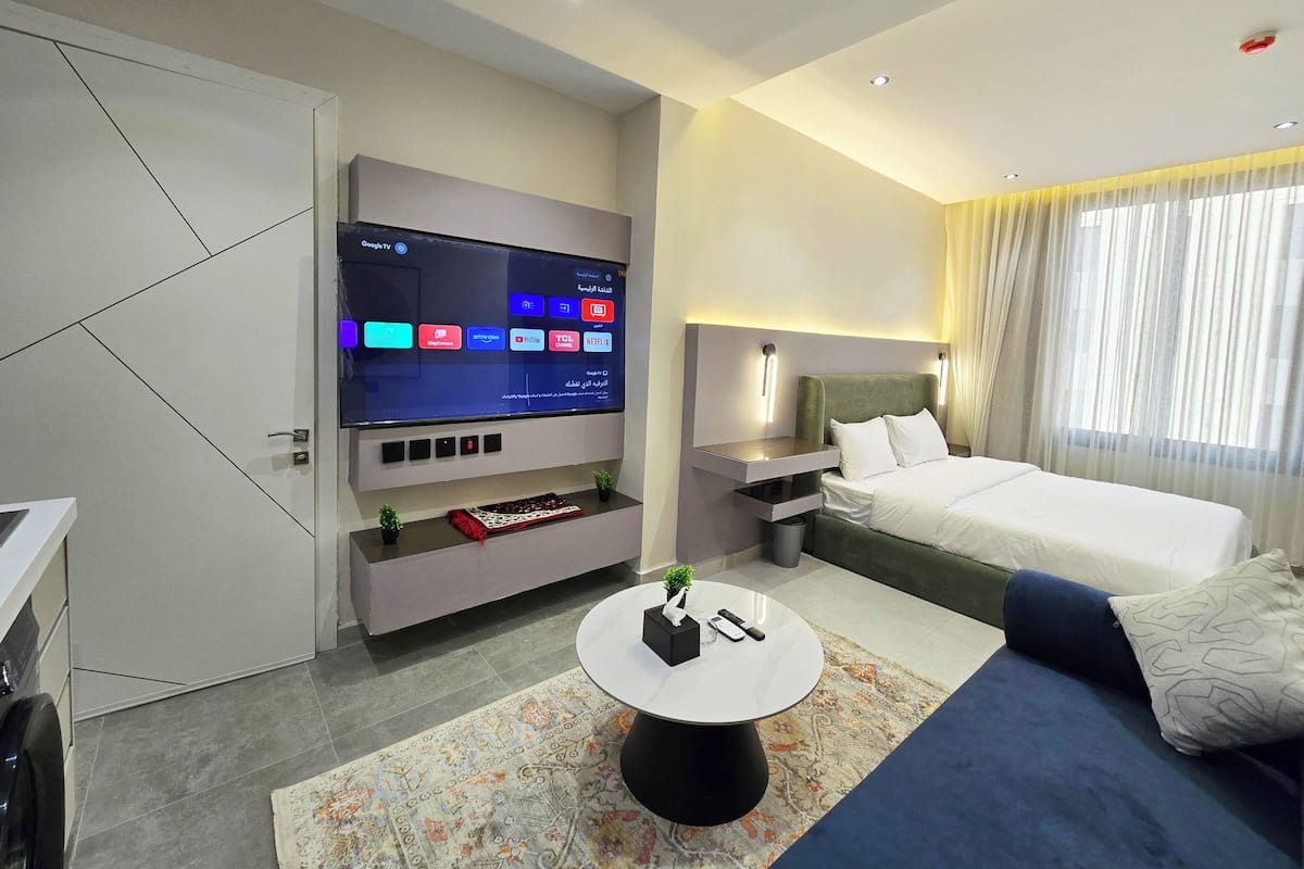 Ein modernes Hotelzimmer mit einem großen Flachbildfernseher an der Wand, einem Bett mit weißer Bettwäsche, einem kleinen runden Couchtisch und einem Sofa mit blauen Kissen.
