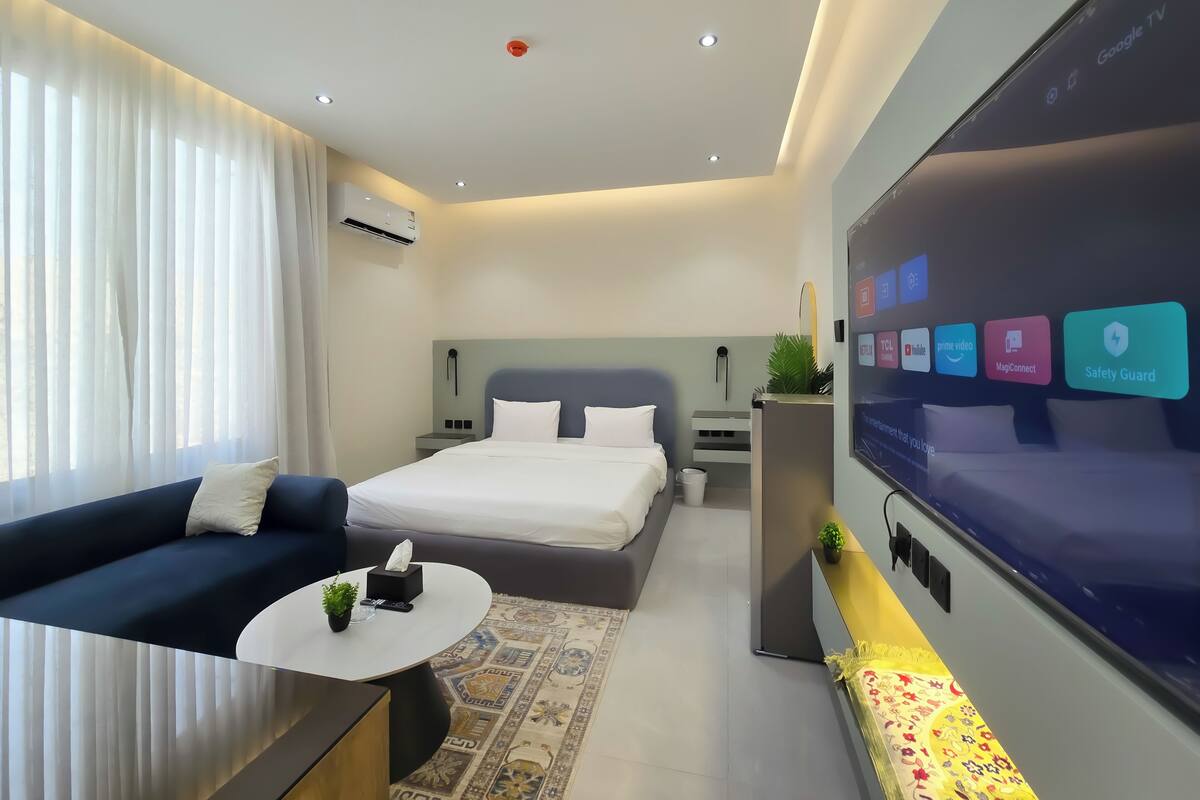 Ein modernes Hotelzimmer mit Bett, Sofa, Fernseher und verschiedenen App-Symbolen sowie einem kleinen Tisch mit einer Pflanze.