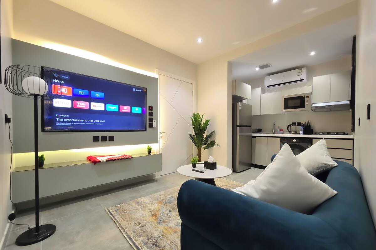 Ein modernes Wohnzimmer mit einem an der Wand befestigten Flachbildfernseher, einem blauen Sofa, einem kleinen Couchtisch und einem gemusterten Teppich.