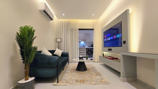 A modern living room with a sofa, a flat-screen TV, a potted plant, and a dining area with a table and chairs. - Hometel NRJS B Ruh (Riyadh)