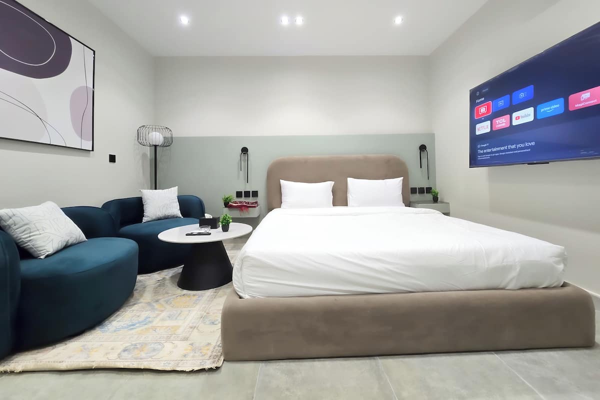 Ein modernes Hotelzimmer mit einem Bett, zwei Sofas, einem Couchtisch und einem Fernseher, der Streaming-Dienste anzeigt.