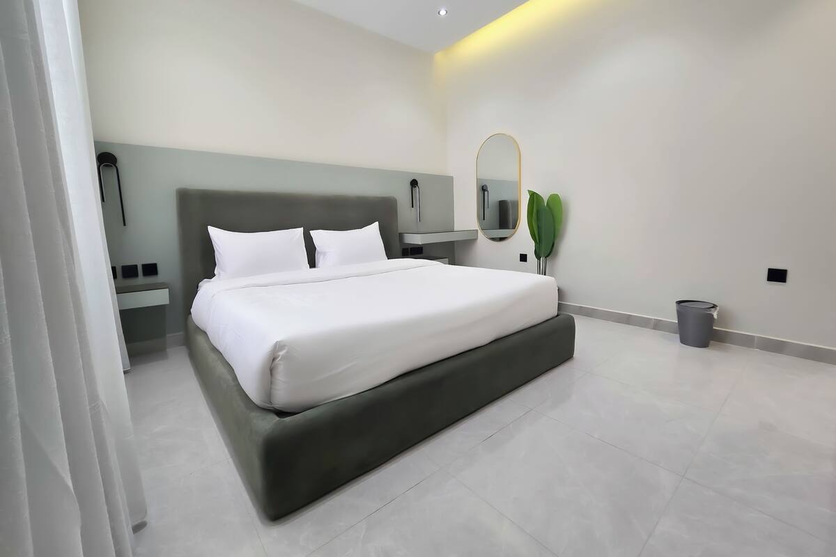 Ein modernes Schlafzimmer mit einem großen Bett, einem Nachttisch, einem wandmontierten Spiegel und einer kleinen Mülltonne.