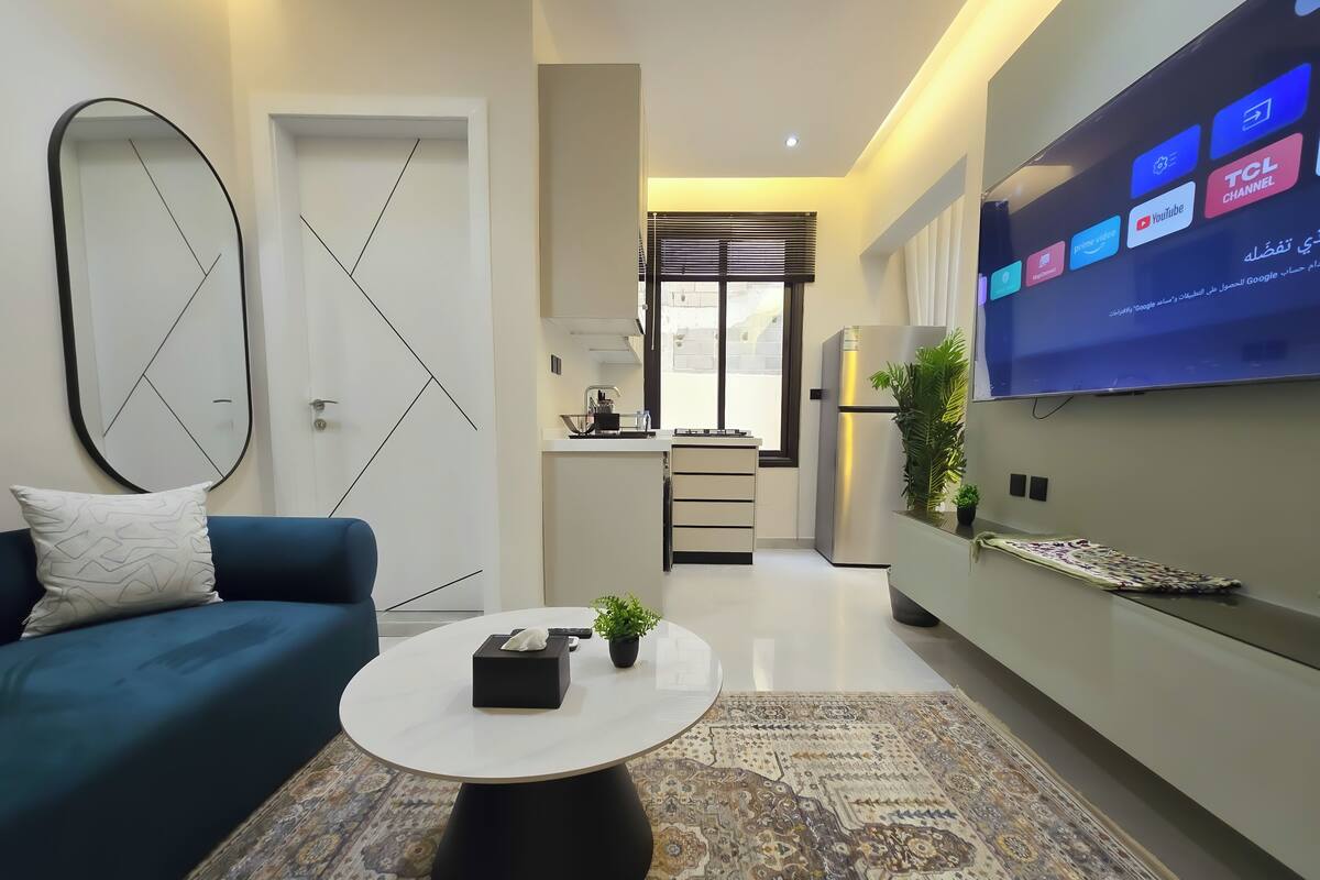 Ein modernes Wohnzimmer mit einer Couch, einem Couchtisch und einem gemusterten Teppich. Im Hintergrund ist ein Küchenbereich mit einem Kühlschrank und einem Fernseher, der verschiedene Sender zeigt, erkennbar.