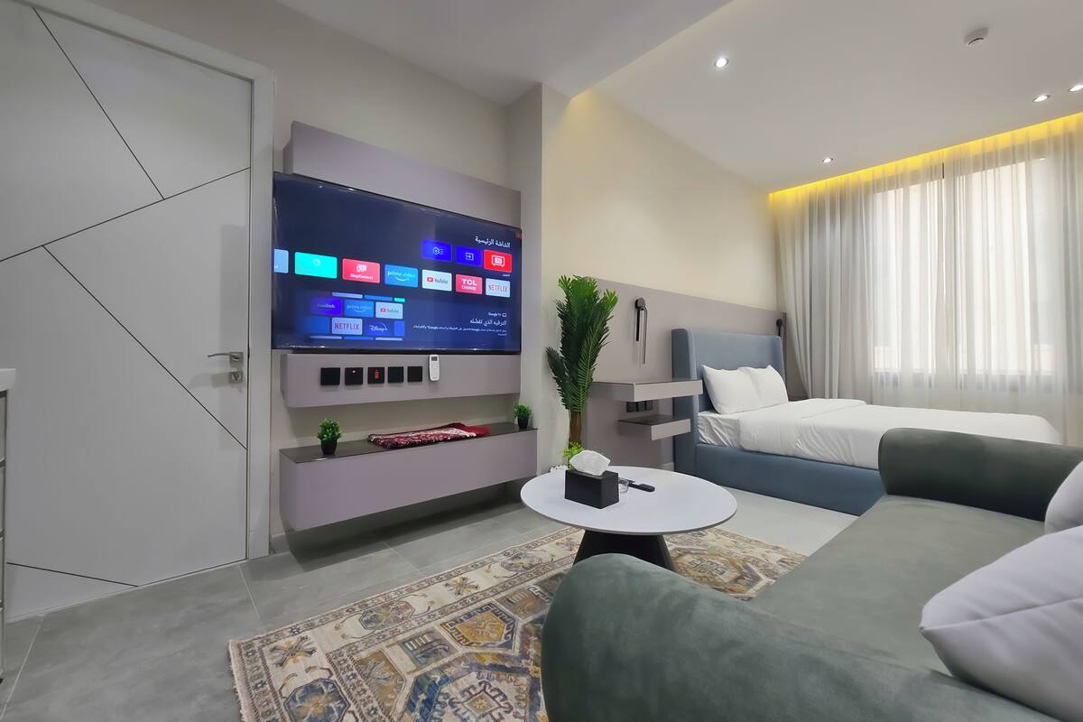 Ein modernes Hotelzimmer mit Flachbildfernseher, der Streaming-Dienste anzeigt, einem Bett mit Kopfteil, einer Couch, einem kleinen Tisch und einem dekorativen Teppich.