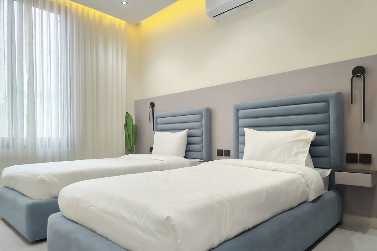 Ein modernes Hotelzimmer mit zwei Betten, einem großen Fenster mit durchscheinenden Vorhängen, einer Wand-Klimaanlage und einem Nachttisch mit Lampe.