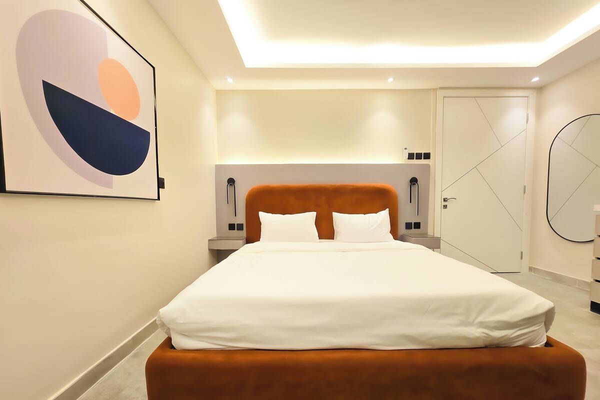 Ein modernes Hotelzimmer mit einem großen Bett, einem geometrisch gestalteten Kopfteil, einem Nachttisch, einem wandmontierten Spiegel und einem abstrakten Kunstwerk.