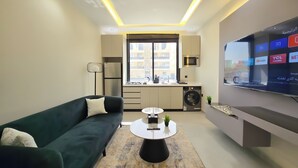 A modern living room with a sofa, a coffee table, and a TV. There is a kitchen area with a microwave and a washing machine. - Hometel NRJS B Ruh (Riyadh)