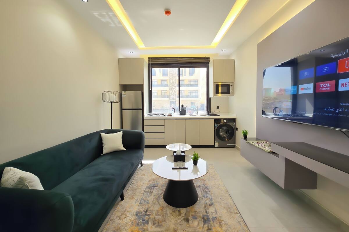 Ein modernes Wohnzimmer mit einer Couch, einem Couchtisch und einem Fernseher. Es gibt eine Küchenzeile mit einer Mikrowelle und einer Waschmaschine.