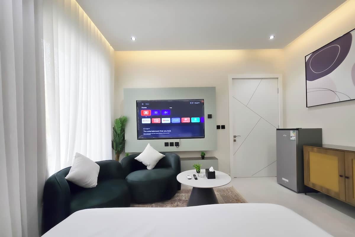 Ein modernes Hotelzimmer mit einem großen Bett, einer Sitzecke mit Couchtisch, einem Flachbildfernseher, der einen Startbildschirm zeigt, und einem Kühlschrank.
