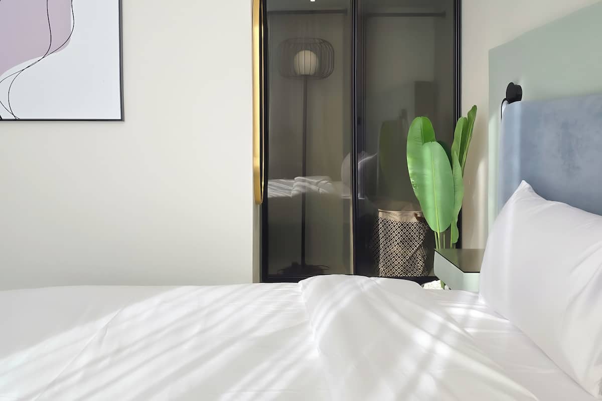 Ein Hotelzimmer mit Bett, Wand-Klimaanlage, gerahmtem abstrakten Kunstwerk, Schiebetürenschrank, Zimmerpflanze und Kopfteil.