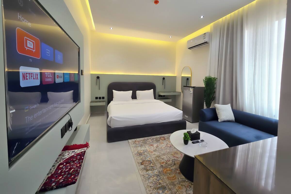 Ein modernes Hotelzimmer mit einem großen Bett, einer Sitzecke, einem Fernseher, der Unterhaltungsangebote zeigt, und einem kleinen Tisch mit einer Pflanze.