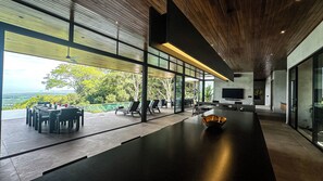 A modern interior with a long dining table, chairs, a TV, and a view of the outdoors.