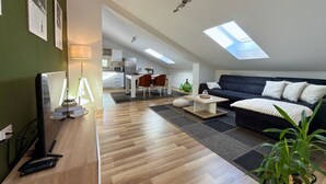 Un appartement moderne de style « loft », comprenant un coin repas, un salon avec un canapé et une télévision, et une cuisine située à l’arrière-plan.