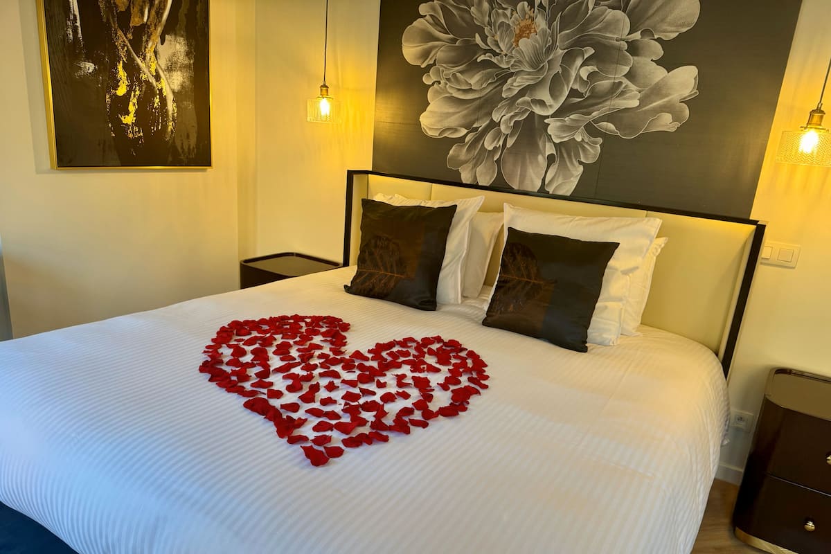 Une chambre d’hôtel avec un grand lit, une composition en forme de cœur faite de pétales de roses sur le lit, un grand tableau floral accroché au mur et deux lampes de chevet.