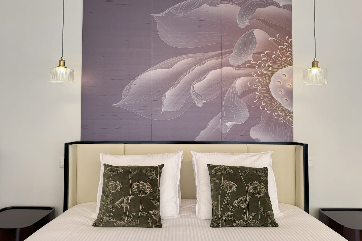 Une chambre à coucher avec une grande œuvre d’art florale au mur, deux lampes de chevet et deux coussins ornés de motifs floraux.