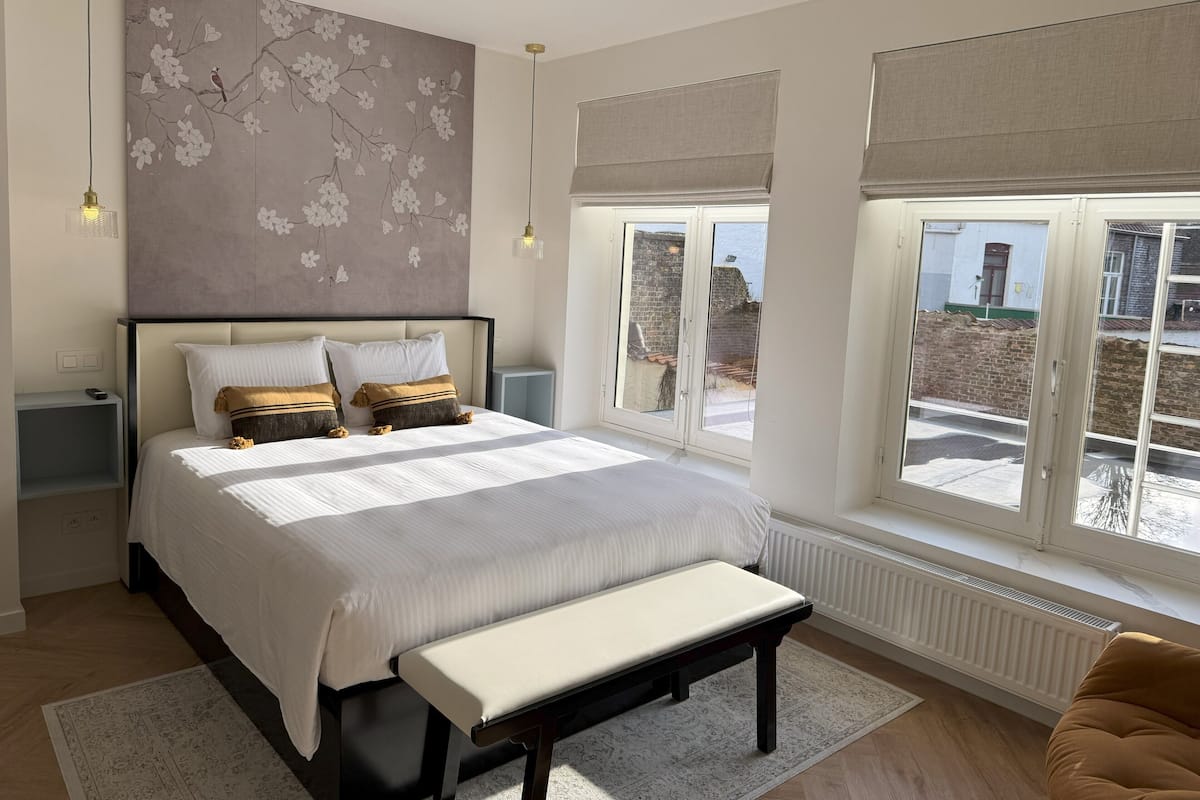 Une chambre avec un grand lit, un banc, un mur décoré de motifs floraux et une vue sur l’extérieur.