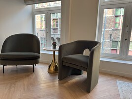 A modern living room with two dark armchairs, a small round table with a gold base, and a view of a brick building through large windows.