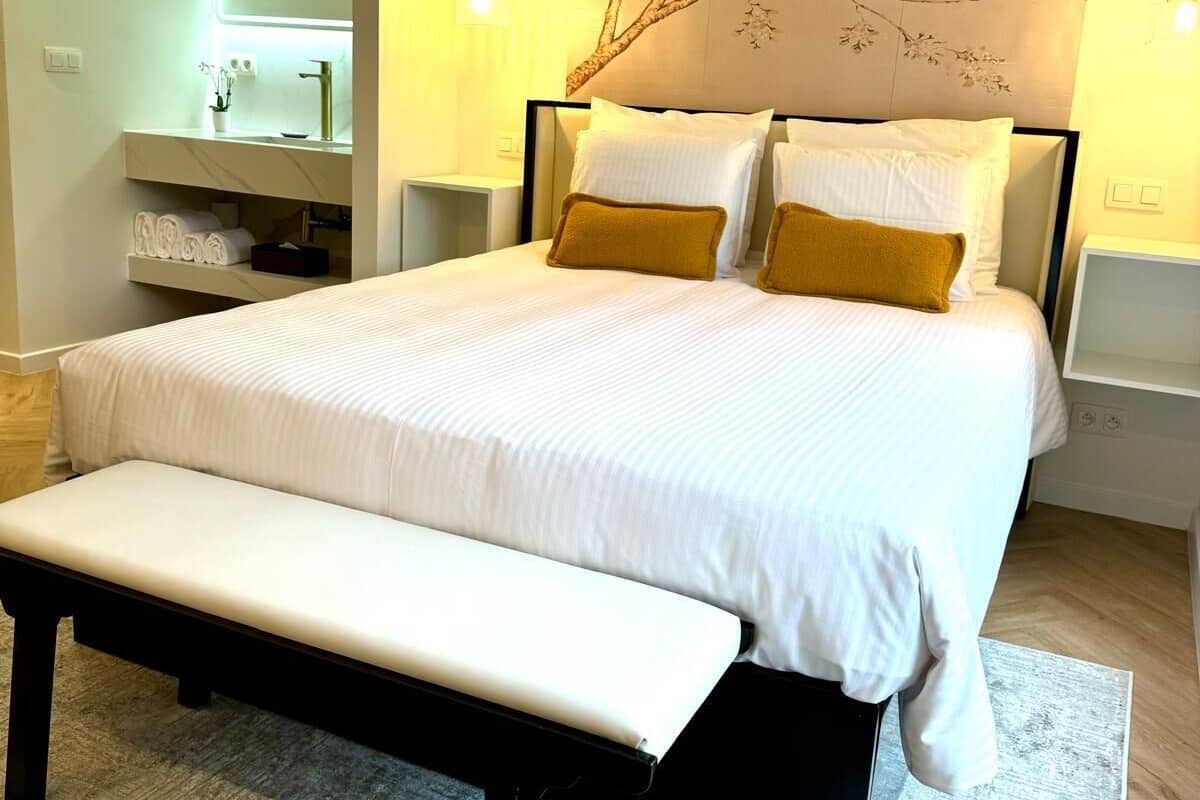Une chambre à coucher moderne avec un grand lit, des tables de chevet, un banc et une œuvre d’art accrochée au mur.
