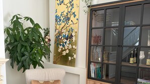 Een moderne woonkamer met een lichtgekleurde fauteuil, een grote potplant, een decoratief schilderij en een houten kast met glazen deuren.